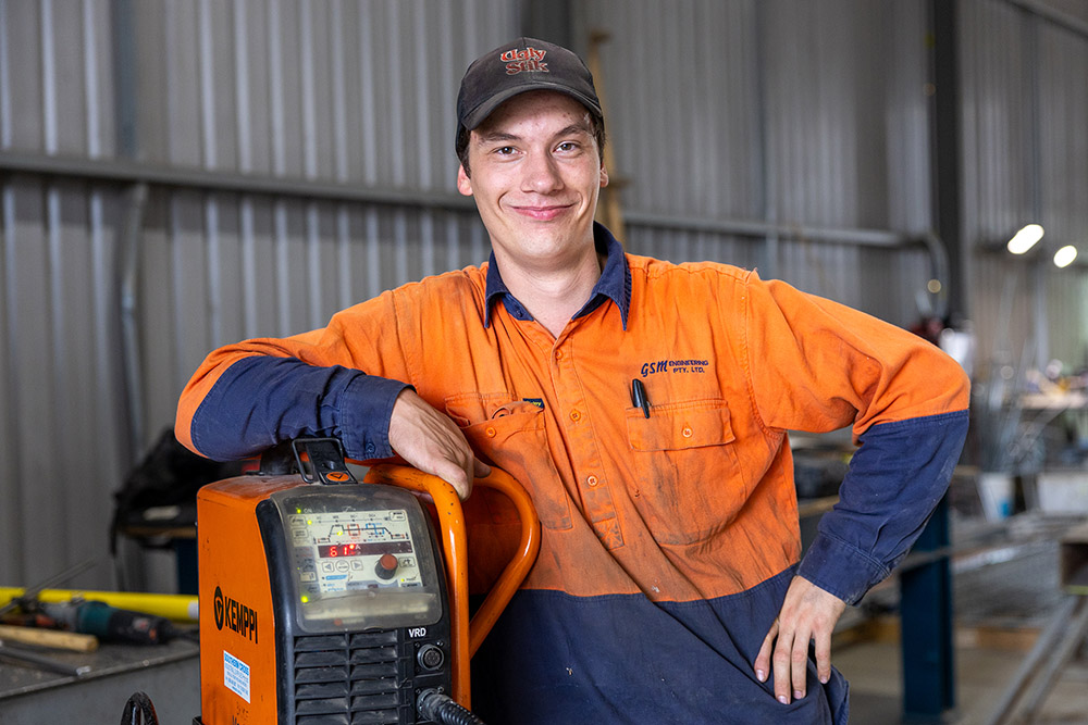 Young Male Engineering Apprentice smiling whilst leaning against welding equipment