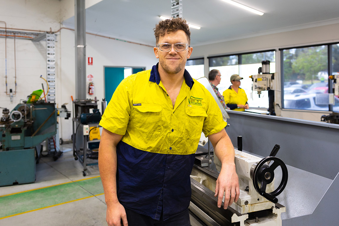 Mature aged male apprentice studying and working in engineering - mechanical trade. They are in front of a lathe, wearing their employers work gear.